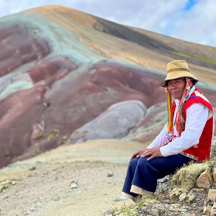 A large rainbow mountain with rocks, some shrubs, and a tourist.