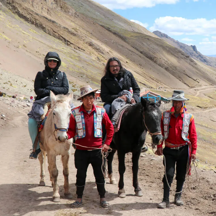 Two tourists ride horses led by local guides in traditional attire, following a rugged trail with Andean mountains rising in the background under a bright sky.