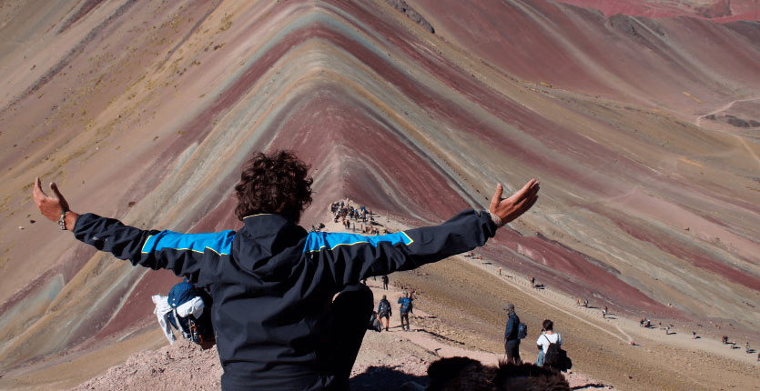 A man with his arms spread out on both sides stands in front of the Rainbow Mountains of Peru, with several people shown moving ahead in the background.