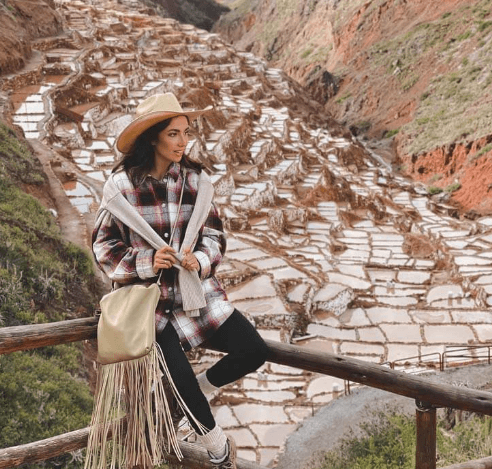 cusco sacred valley tour sacred valley A stylish woman sits on a wooden railing overlooking the Maras Salt Mines in Peru, dressed in layered clothing and a wide-brimmed hat.