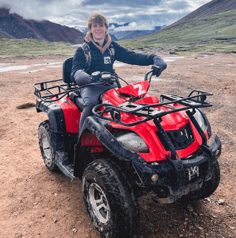 A young traveler smiles while sitting on a red ATV, ready for adventure against the dramatic backdrop of rugged Rainbow Mountain terrain and cloudy skies.