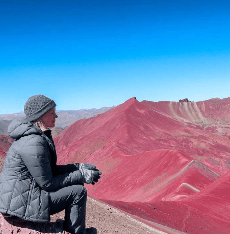 A woman in winter clothing sits peacefully on a rocky ledge, gazing out at the striking crimson ridges and vibrant landscapes of Peru’s Red Valley under a clear blue sky.