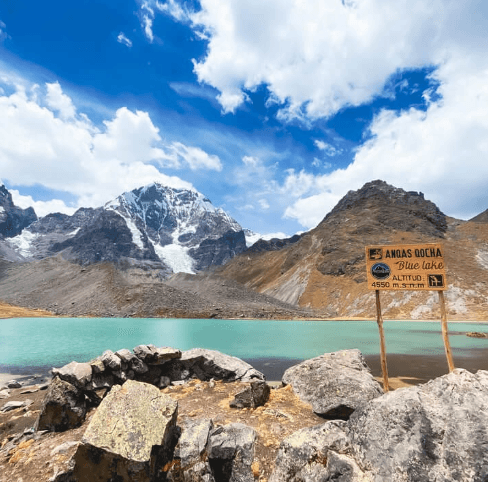 A vibrant turquoise lake, known as Blue Lake, sits beneath snow-capped peaks in the Andes, with a wooden sign marking an altitude of 4550 meters.