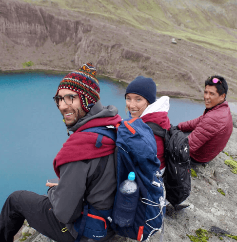 Three smiling hikers in warm gear sit beside a vibrant turquoise lake, backpacks ready, along the rugged Ausangate Trek in Peru.