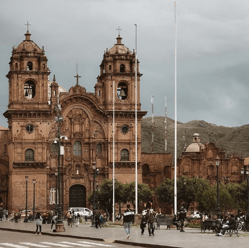 A wide-angle view of the baroque-style Cusco Cathedral with twin bell towers, surrounded by people walking and relaxing in the main plaza, with Andean hills in the background.