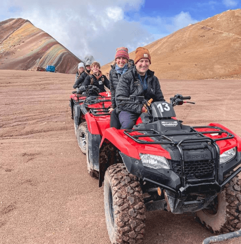 A group of smiling travelers seated on red ATVs lined up on a rugged dirt path, ready for an adventurous ride near Rainbow Mountain in Peru.
