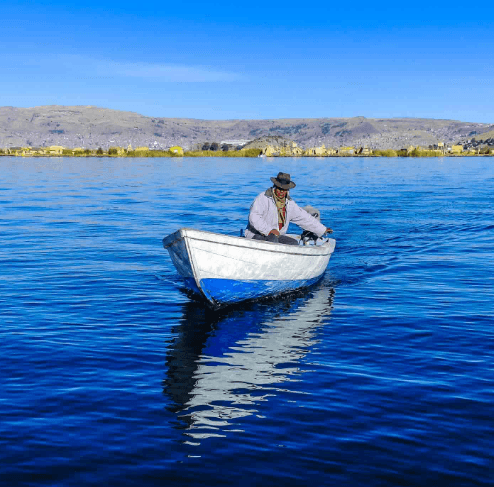 A man wearing traditional Andean clothing rows a small white and blue boat across the deep blue waters of Lake Titicaca, with Puno's shoreline in the distance.