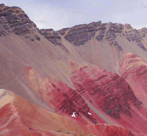 A panoramic view of Peru’s Red Valley with striking layers of red, maroon, and tan mountains under a cloudy sky, showcasing natural mineral formations.