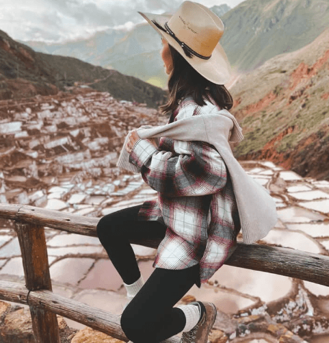 A woman in a plaid shirt and wide-brimmed hat poses on a wooden railing overlooking the salt terraces of Maras in Peru’s Sacred Valley.