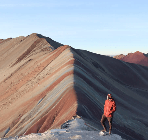 A traveler in a red jacket stands near the summit of Rainbow Mountain at sunrise, gazing at the colorful striped ridges under a clear blue sky.