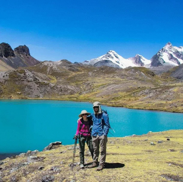 A couple stands beside a turquoise alpine lake, surrounded by rugged mountain peaks and clear blue skies during a two-day Rainbow Mountain trek.