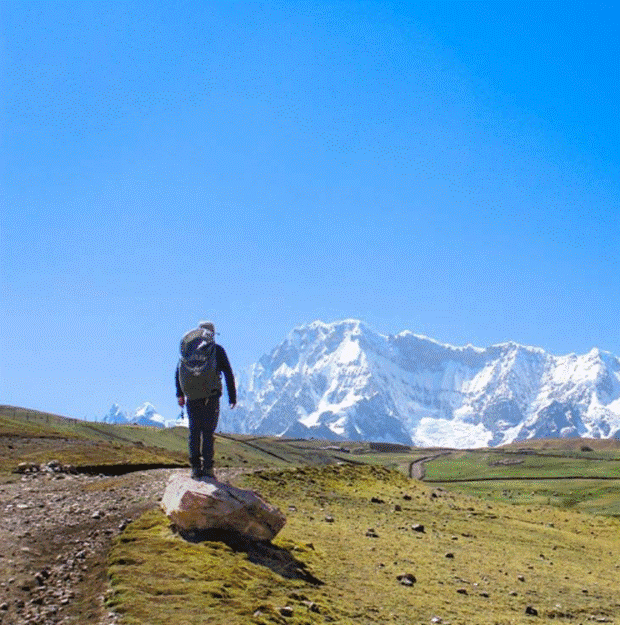 A hiker walking on a dirt trail toward snow-covered peaks of Ausangate under a clear blue sky.