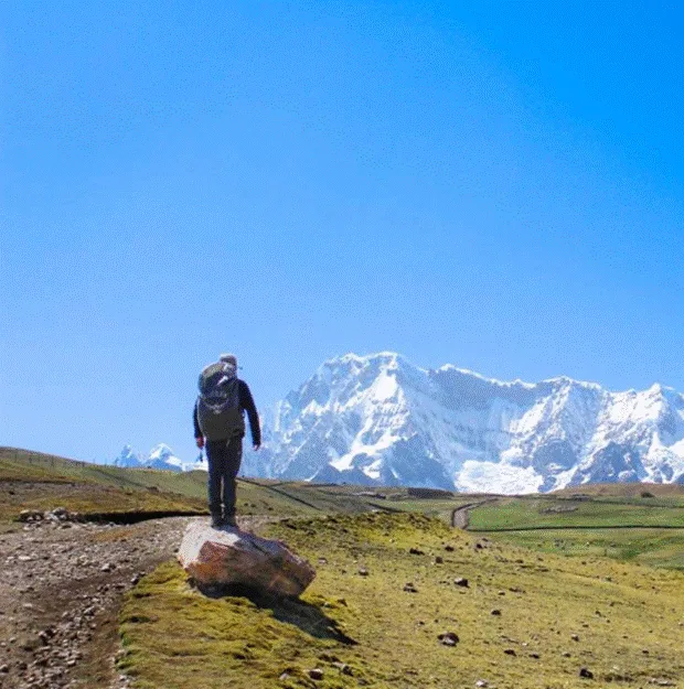 A hiker walking on a dirt trail toward snow-covered peaks of Ausangate under a clear blue sky.