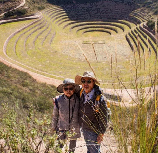 Two travelers in sun hats and sunglasses stand close together along a grassy trail in the Sacred Valley, surrounded by tall green reeds and golden sunlight.