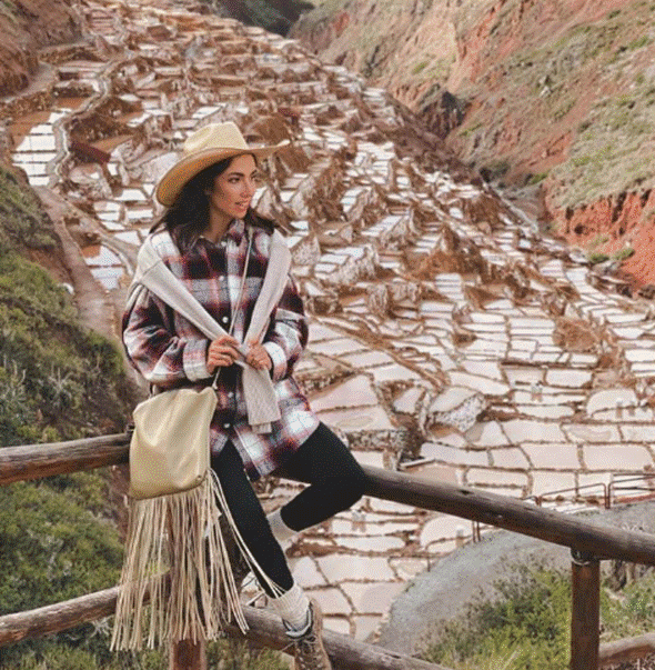 A woman in a plaid shirt, scarf, and hat sits on a wooden railing with the terraced salt ponds of Maras in the Sacred Valley spread out behind her.