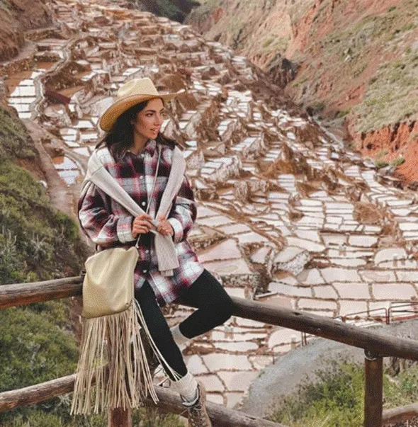 A woman in a plaid shirt, scarf, and hat sits on a wooden railing with the terraced salt ponds of Maras in the Sacred Valley spread out behind her.