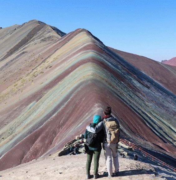 A couple stands arm in arm at the base of the striped Rainbow Mountain in Peru, gazing up at its colorful mineral bands under a clear blue sky.