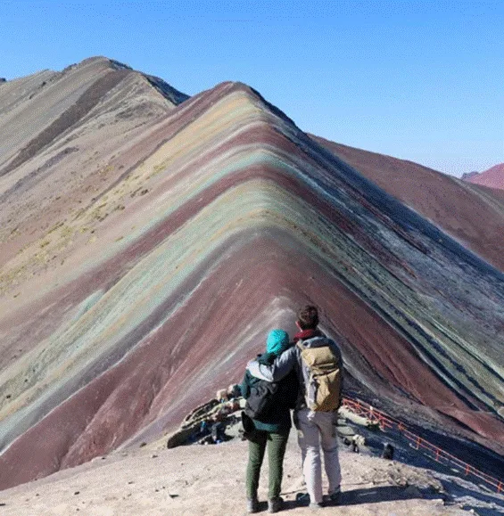 A couple stands arm in arm at the base of the striped Rainbow Mountain in Peru, gazing up at its colorful mineral bands under a clear blue sky.
