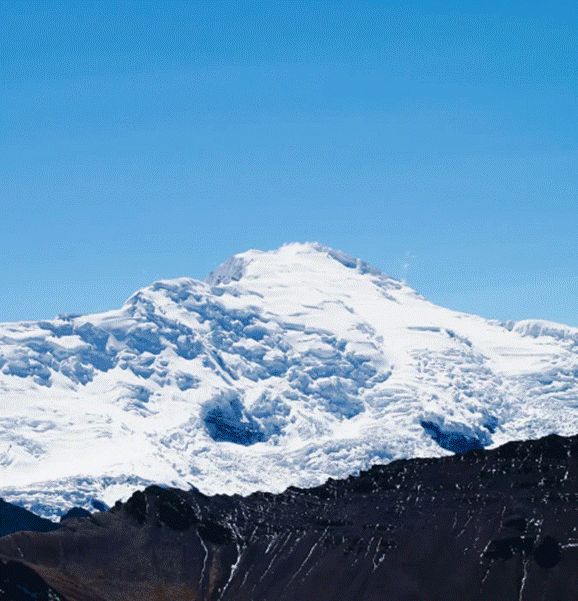 Snow-covered Ausangate mountain peak under a clear blue sky with dark rocky terrain in the foreground.