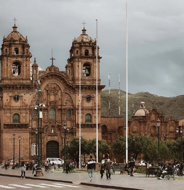 People walking and sitting in front of a historic colonial cathedral with mountains in the background.