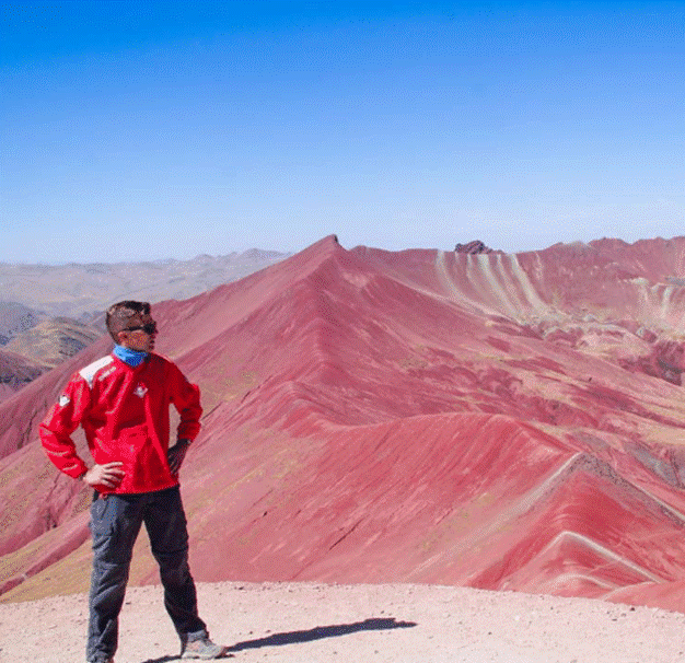 A man in a red jacket standing at the summit with the striking red ridges of Rainbow Mountain and Red Valley behind him.