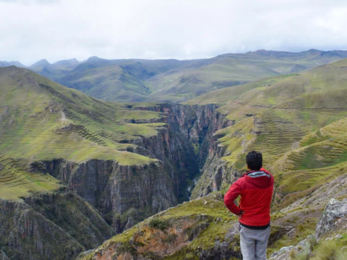 A hiker in a red jacket standing on a rocky ridge overlooking a dramatic, deep canyon.
