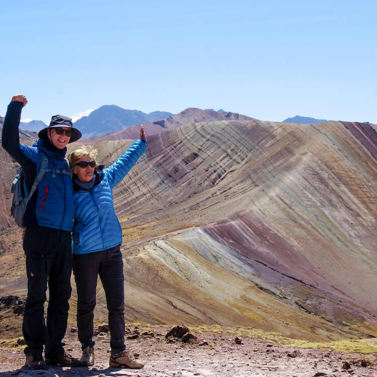Two hikers celebrating with raised arms at the summit of Rainbow Mountain in Peru.