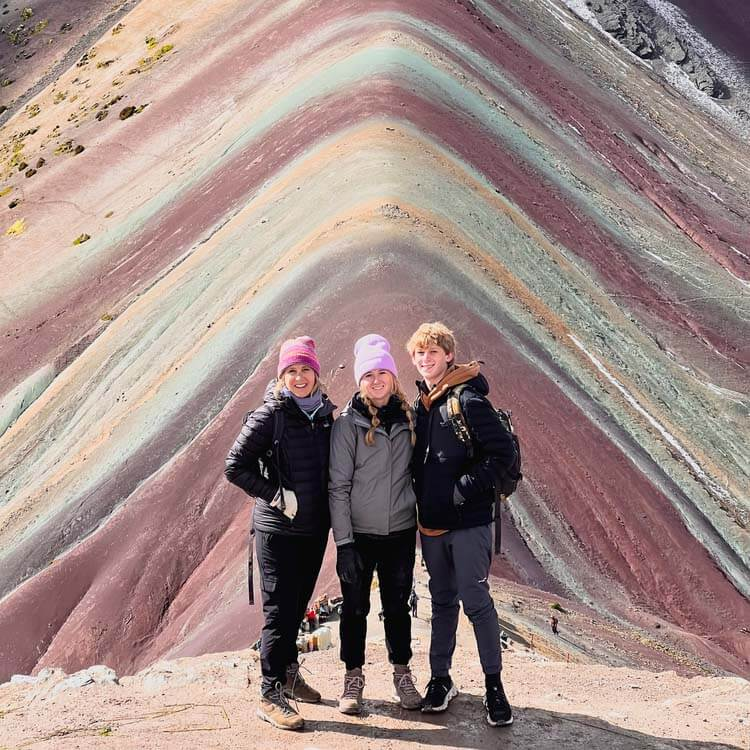 Three smiling hikers standing together in front of the vivid multicolored mineral stripes of Rainbow Mountain.
