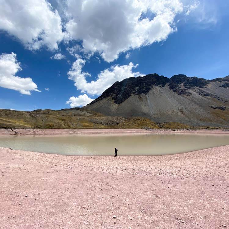 what to eat trekking peru rainbow mountain alpine lake A lone hiker standing beside a still alpine lake on red mineral terrain.